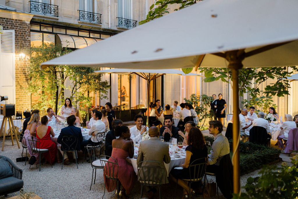 Une grande terrasse extérieure élégante avec plusieurs tables rondes où des groupes de personnes dînent sous des parasols blancs en soirée.
