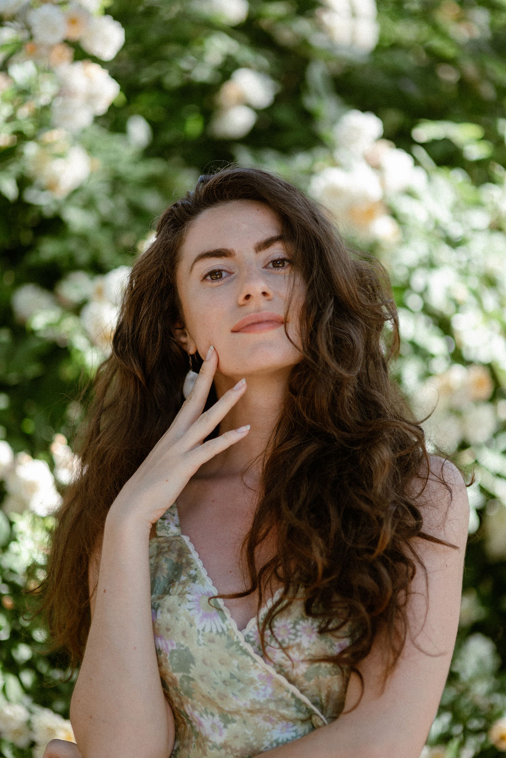 Smiling woman with long wavy brown hair resting her face on her hand against a plain light background.