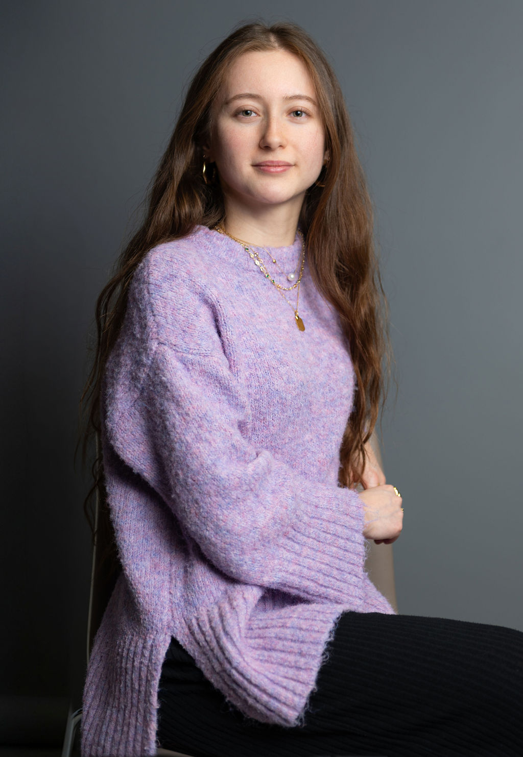 Smiling woman with long wavy brown hair resting her face on her hand against a plain light background.