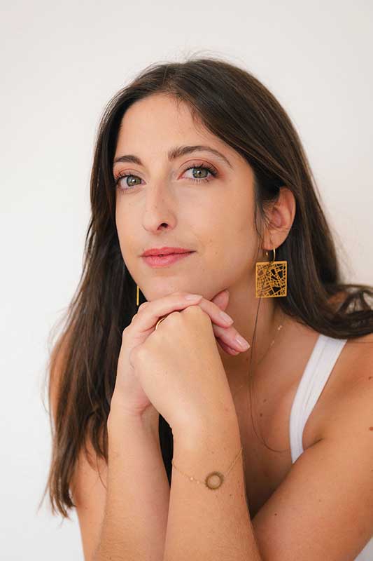 Smiling woman with long wavy brown hair resting her face on her hand against a plain light background.