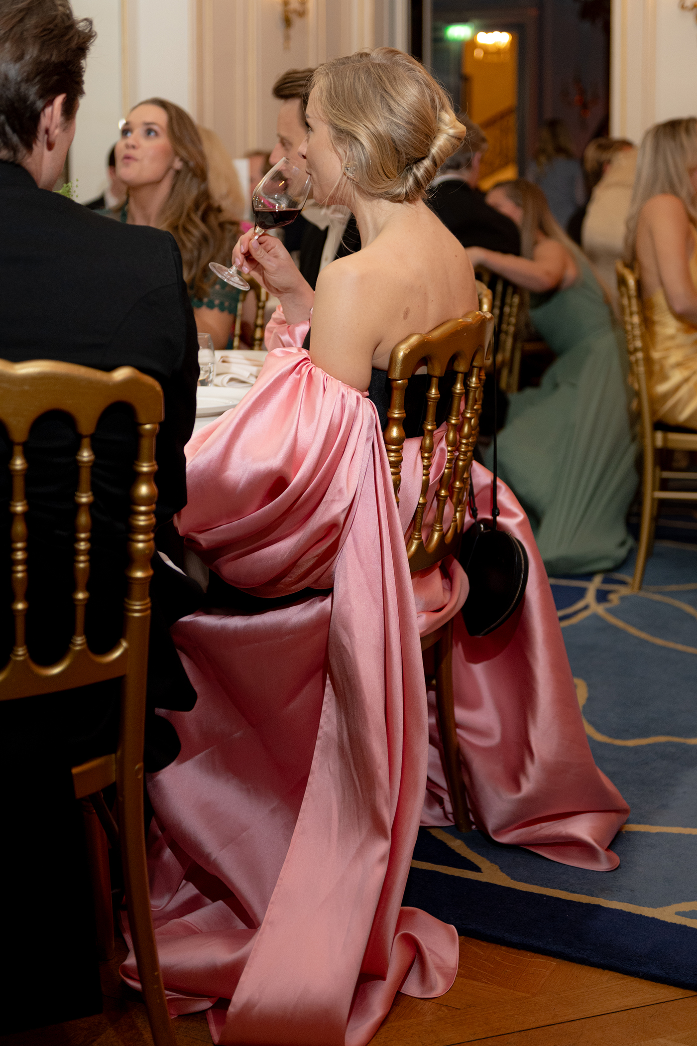Woman in a brown satin dress speaking into a microphone while reading from a piece of paper outdoors next to potted plants.